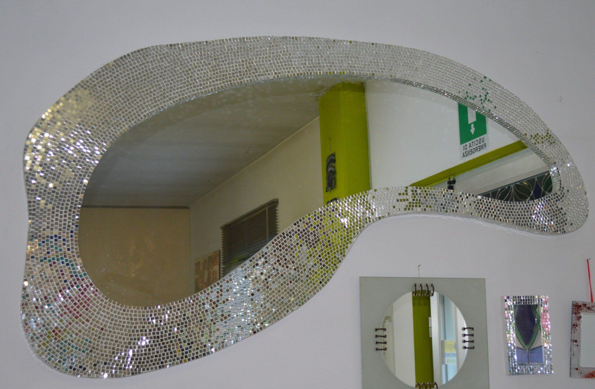 Specchio con cornice in mosaico argentato su parete bianca; riflessi di una stanza con pilastro verde lime.