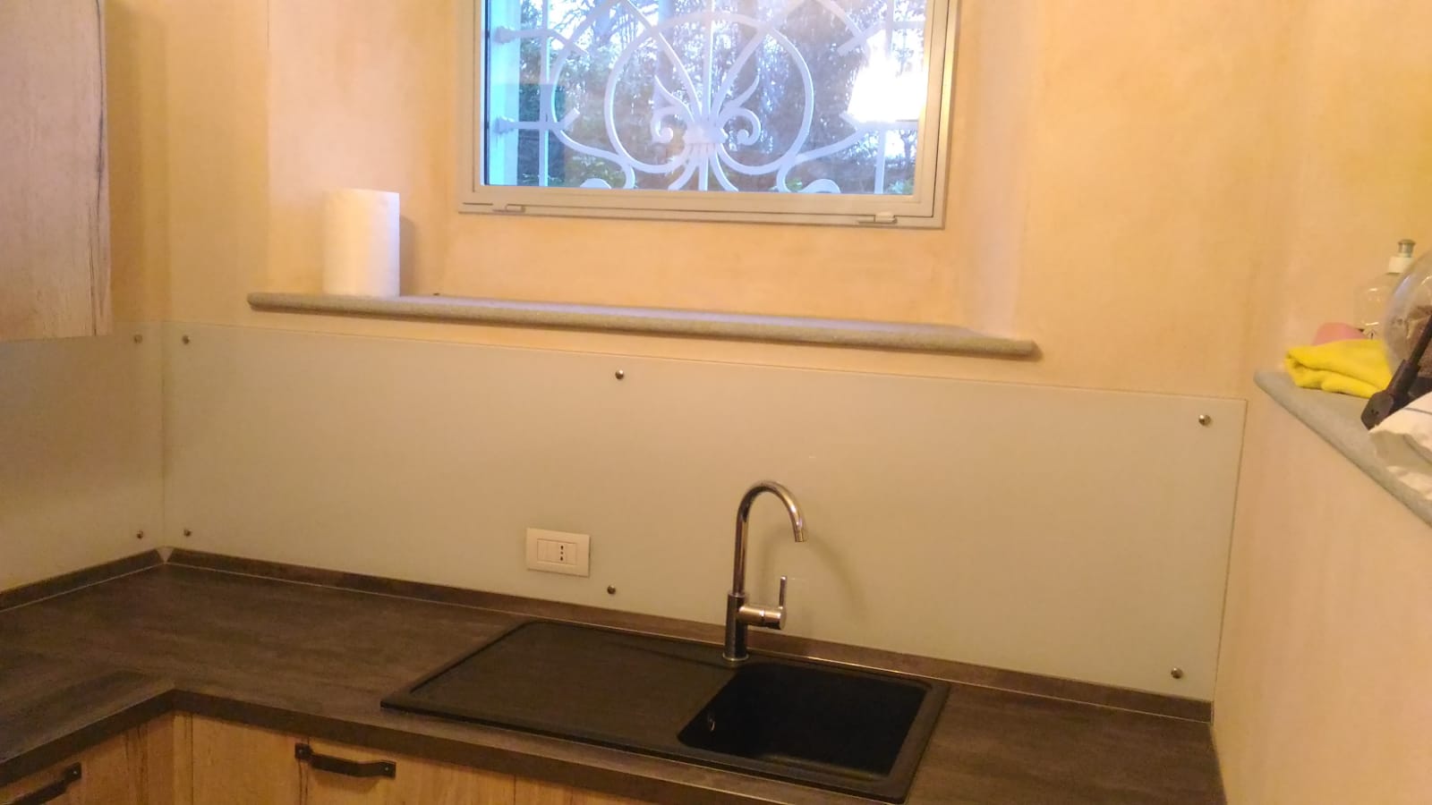 Cucina con lavello e paraschizzi. Piano di lavoro scuro e mobili chiari. Finestra sopra il lavello.