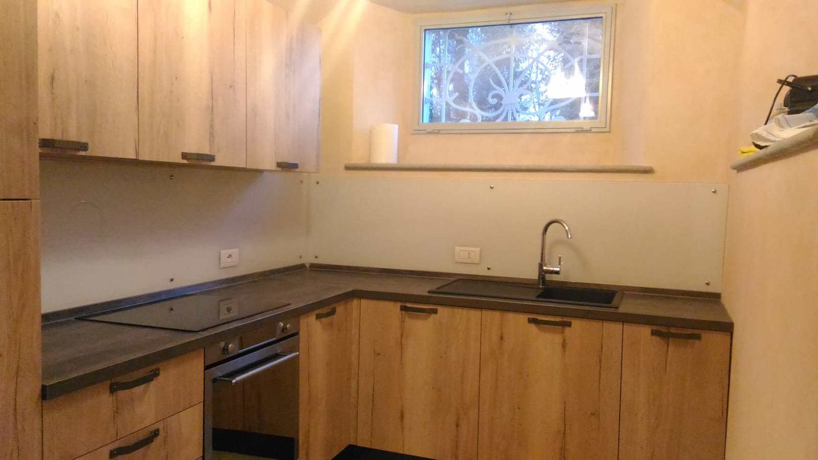 Cucina a L con mobili in legno chiaro, piano di lavoro scuro e un lavandino sotto una piccola finestra.