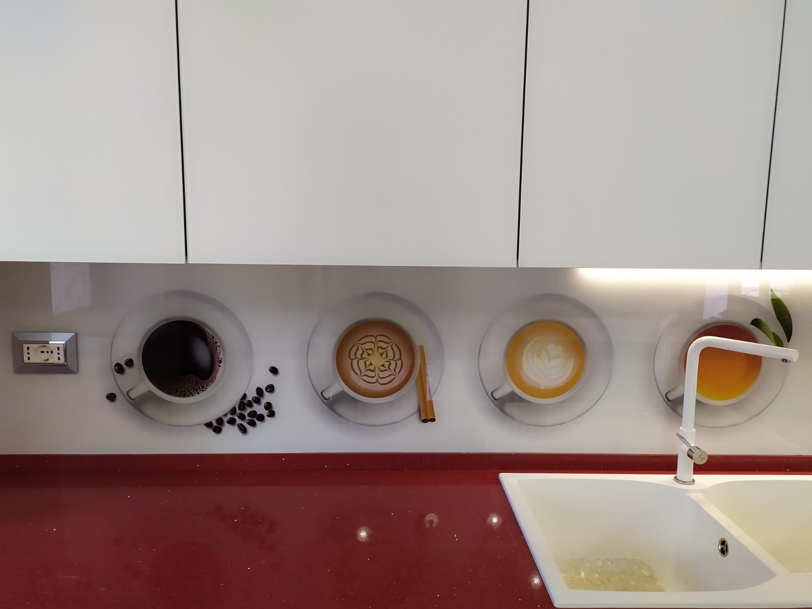 Paraschizzi della cucina con motivi a tazzine da caffè, bancone rosso, mobili bianchi e lavello.
