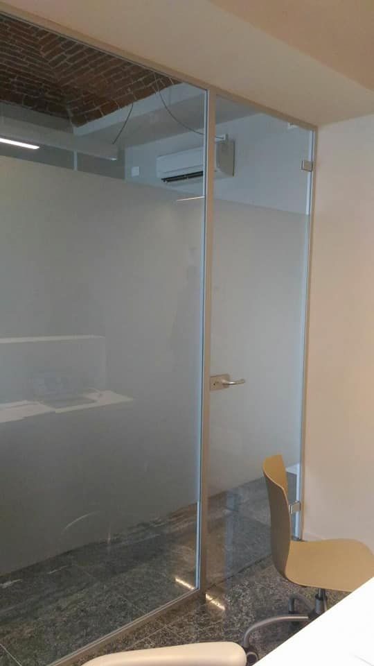 Ufficio in vetro con pareti e porta smerigliate, una sedia e pavimento in piastrelle scure.