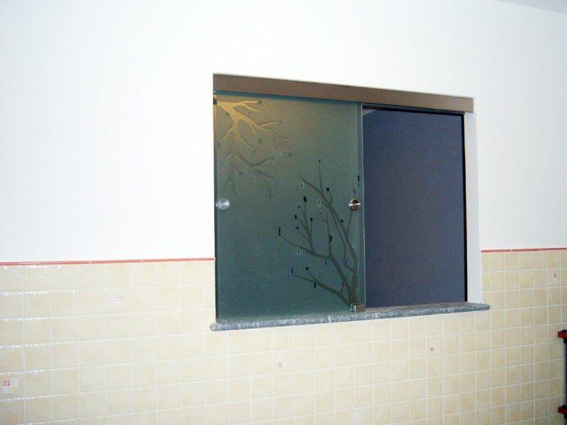 Finestra scorrevole in vetro smerigliato con motivo a rami, sopra un davanzale in granito, inserita in una parete bianca e piastrellata.