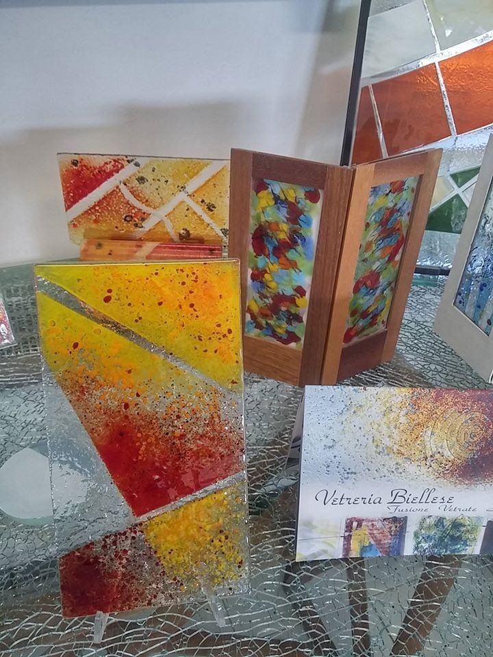 Opere d'arte in vetro colorato, tra cui una lanterna incorniciata e pannelli rettangolari, esposte su una superficie riflettente.