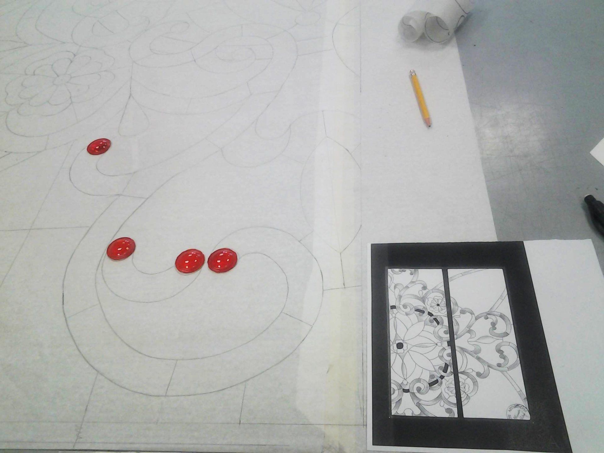 Disegno su un grande foglio di carta con puntini rossi che indicano le posizioni, accompagnato da un disegno di riferimento più piccolo, una matita e un rotolo di carta.