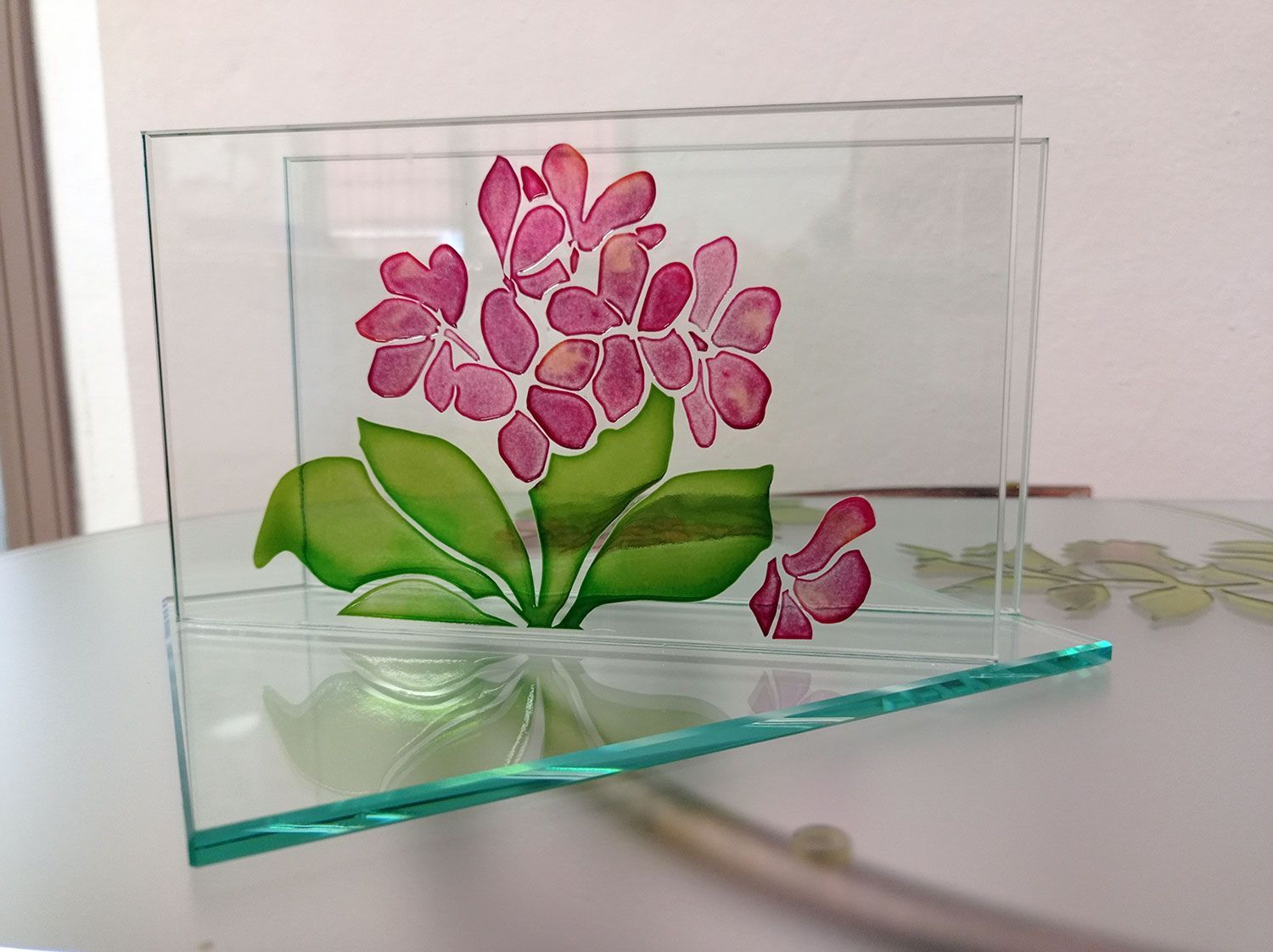 Blocco di vetro con dipinto di fiori rosa e foglie verdi, appoggiato su un tavolo bianco.