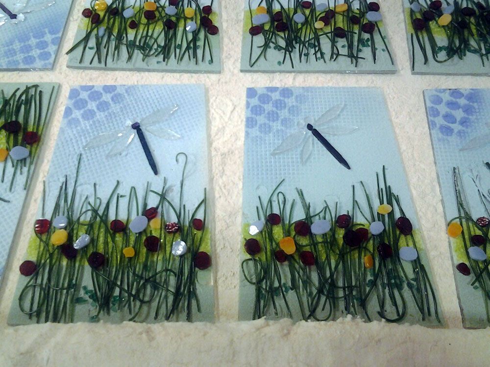 Pannelli di vetro con disegni di libellule e fiori, erba verde, cielo azzurro, vari accenti floreali colorati.