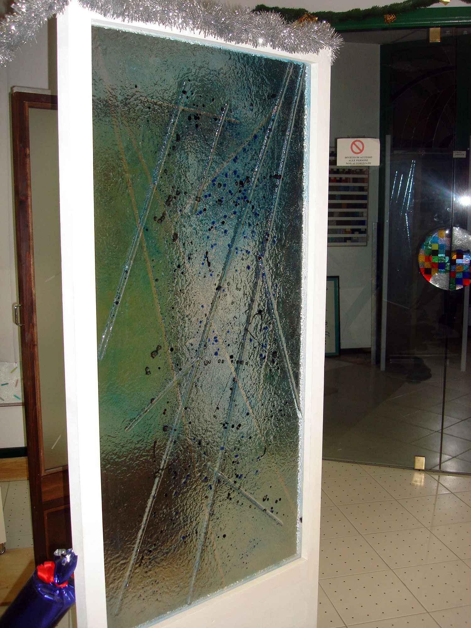 Porta in vetro con cornice bianca e motivo testurizzato, probabilmente piombata. Sullo sfondo si vedono un cartello 