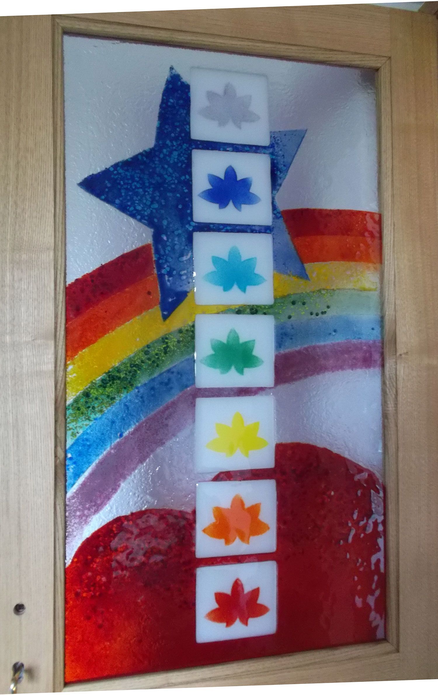 Opera d'arte in vetro colorato: arcobaleno, stella e sette fiori di loto colorati su quadrati bianchi, all'interno di una cornice di legno.