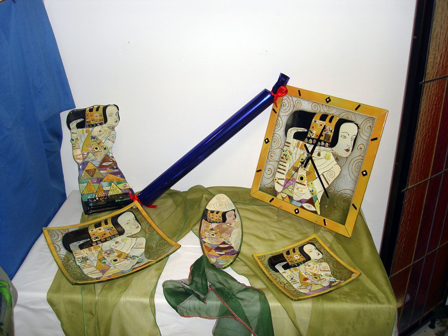 Esposizione di opere d'arte decorative: orologio, piatti, vaso con figure stilizzate su stoffa verde, sfondo blu.