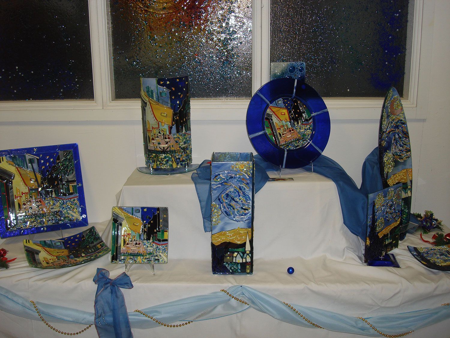 Esposizione di opere d'arte in mosaico, per lo più blu e oro, disposte su un tavolo bianco con tessuto blu.