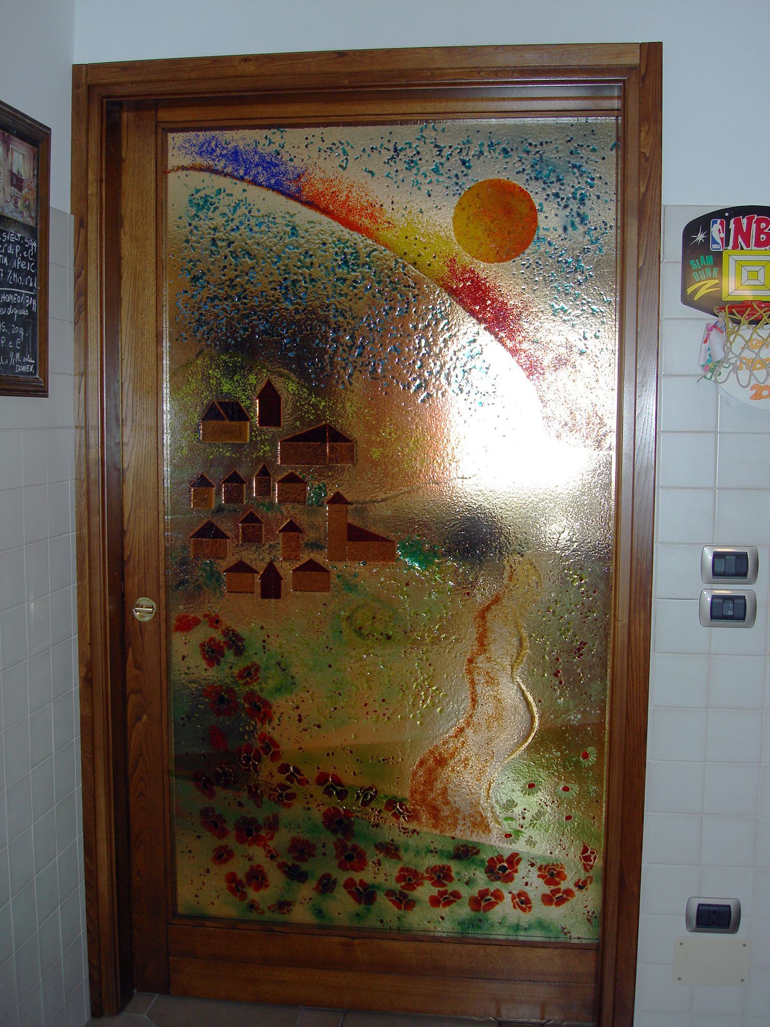 Porta in vetro con un paesaggio colorato dipinto, case e un sole all'interno di una cornice di legno.