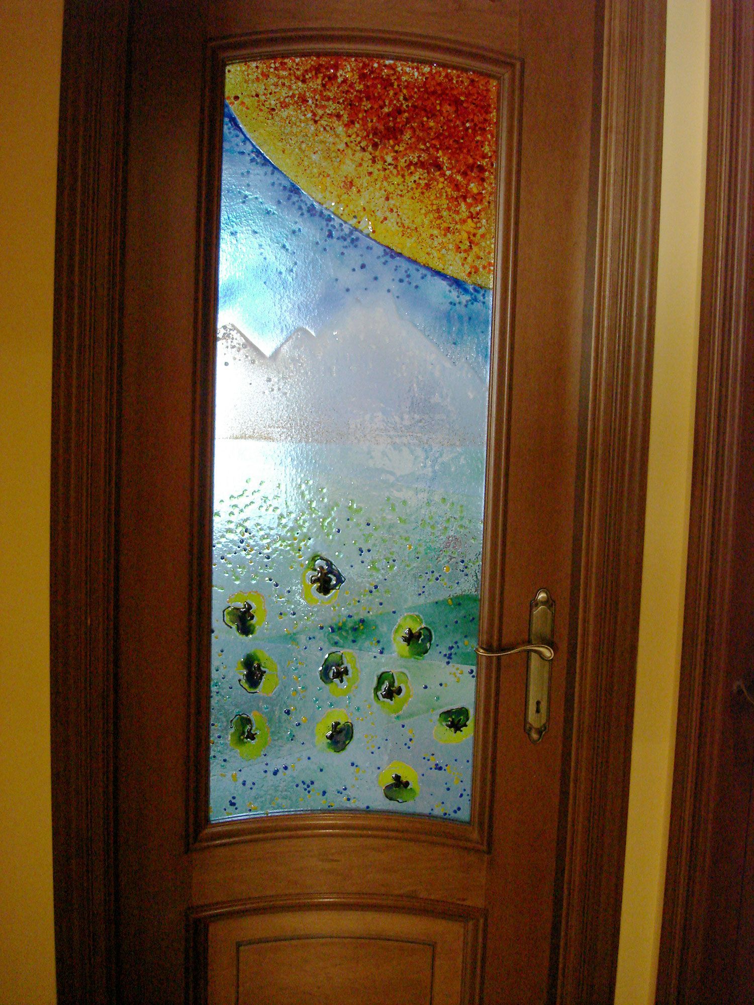 Porta in legno con pannello in vetro colorato raffigurante un paesaggio colorato: sole, montagne e piante verdi e gialle.