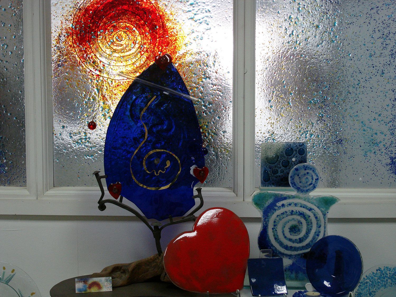 Opere d'arte in vetro blu e rosso esposte su un tavolo davanti a una finestra. Un cuore rosso è in primo piano.