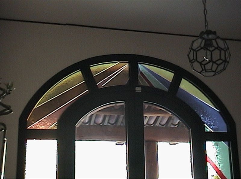 Vetrata ad arco sopra le doppie porte, colori giallo, arancione e blu, finiture nere.