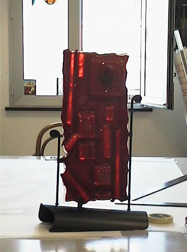Opera d'arte in vetro rosso su un supporto in metallo nero, su un tavolo. Una finestra sfocata sullo sfondo.