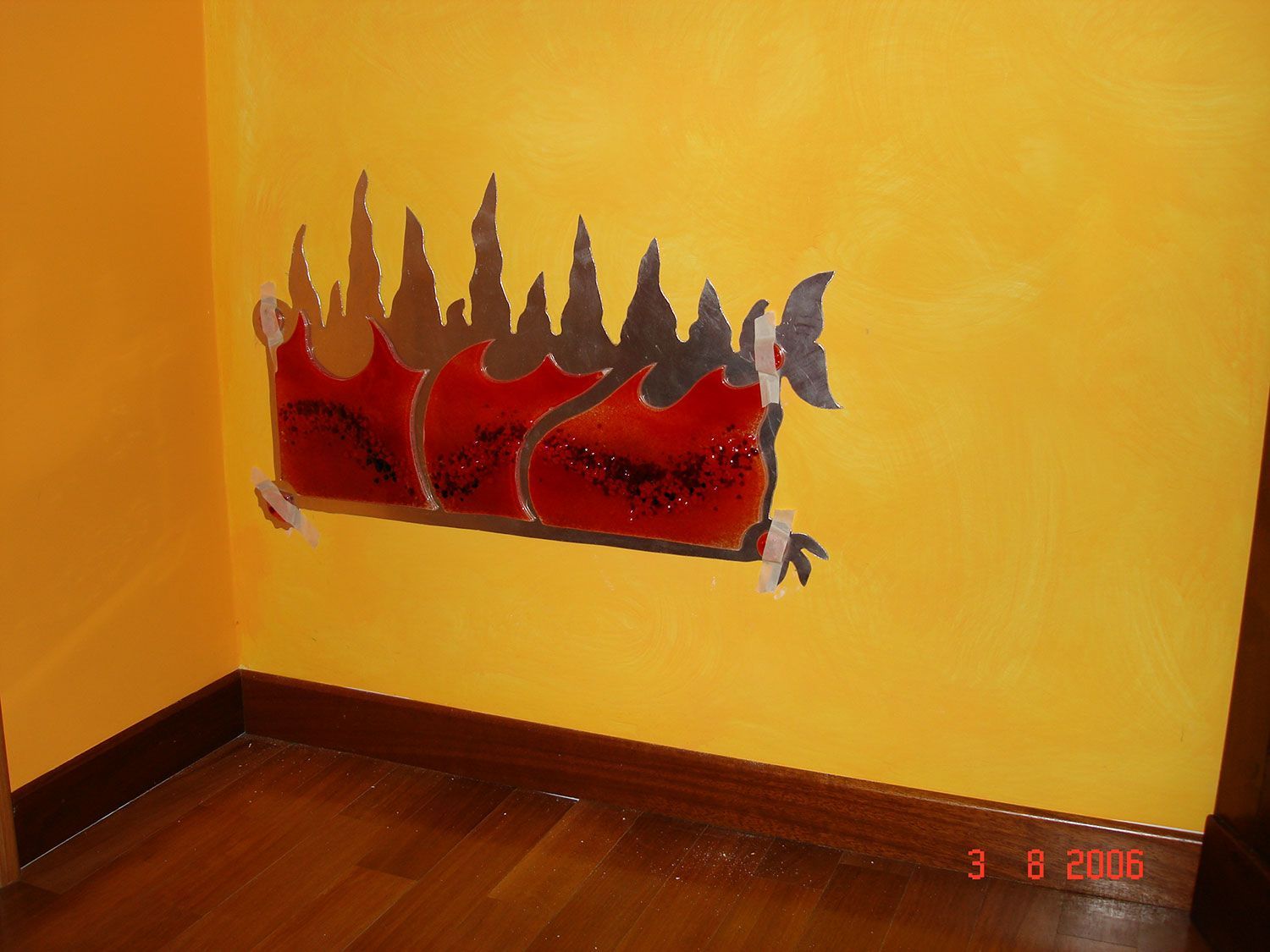 Un'opera d'arte decorativa raffigurante un drago rosso stilizzato in fiamme su una parete gialla e un pavimento in legno marrone.