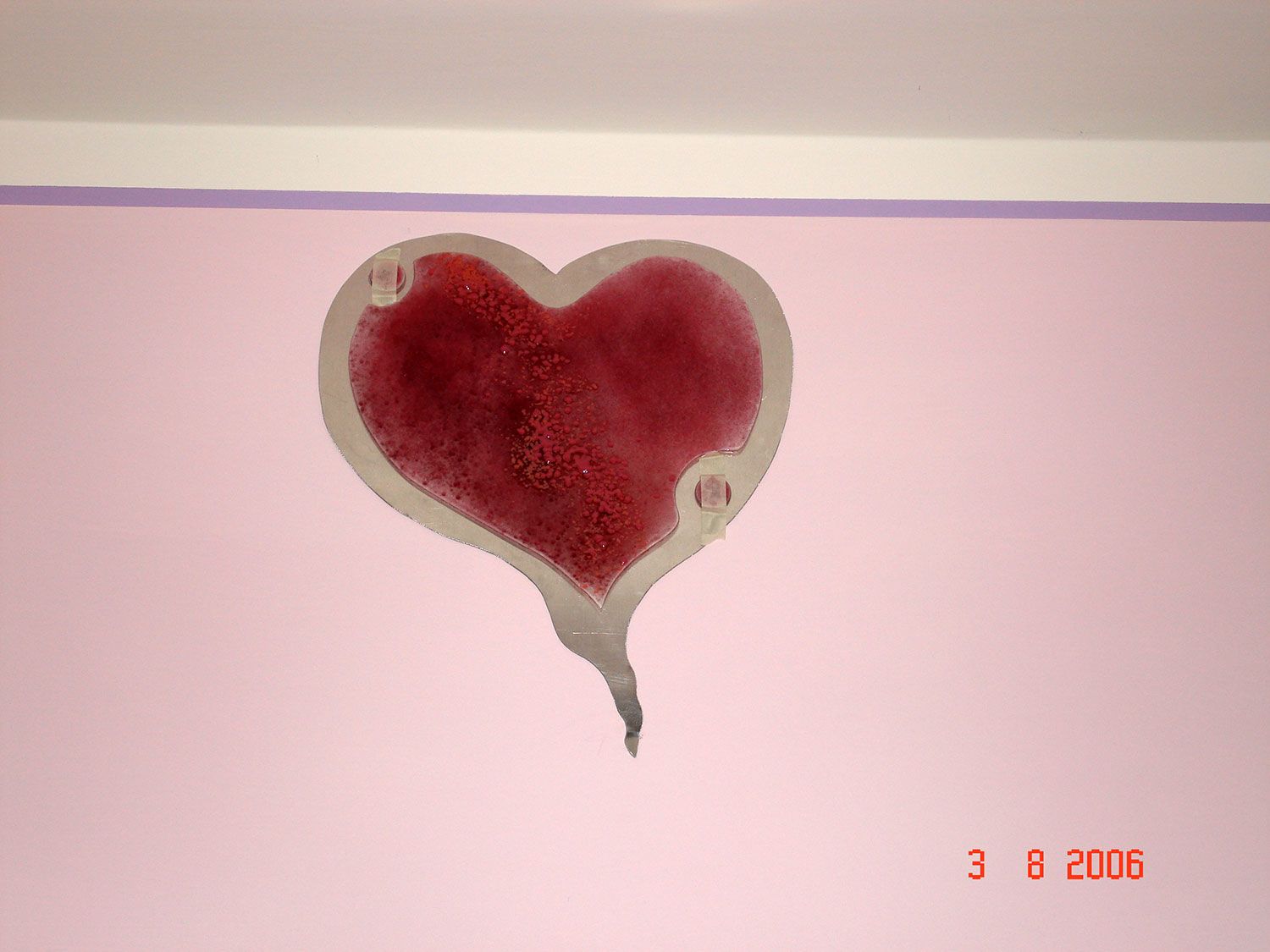 Decorazione a forma di cuore rosso su una parete rosa, con bordo bianco e piccoli fori decorativi. Timbro con data nell'angolo.