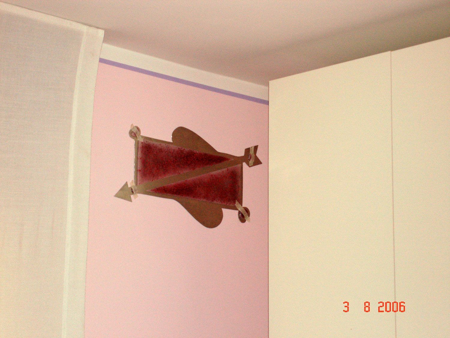 Decorazione a forma di aeroplanino di carta marrone su una parete rosa, con un mobiletto bianco e una tenda sullo sfondo.