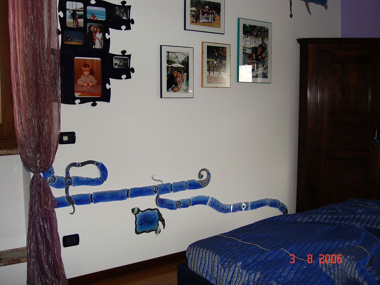 Parete della camera da letto con opere d'arte astratte blu e foto incorniciate, e un armadio in legno scuro.
