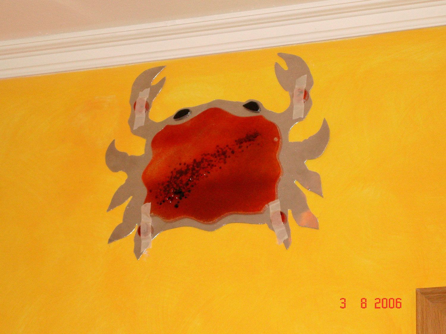 Un ritaglio di granchio rosso su un muro giallo, fissato con nastro adesivo bianco. Il muro ha una rifinitura bianca nella parte superiore.