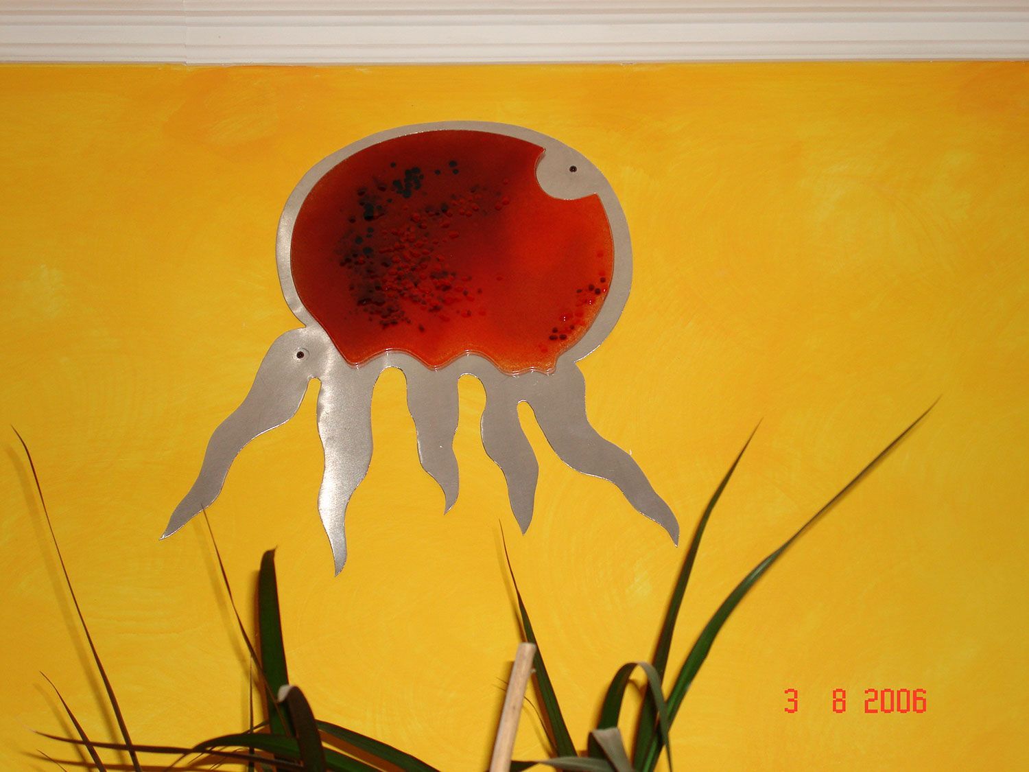 Disegno rosso simile a una medusa su sfondo giallo, con lunghi tentacoli bianchi e alcune piante verdi.