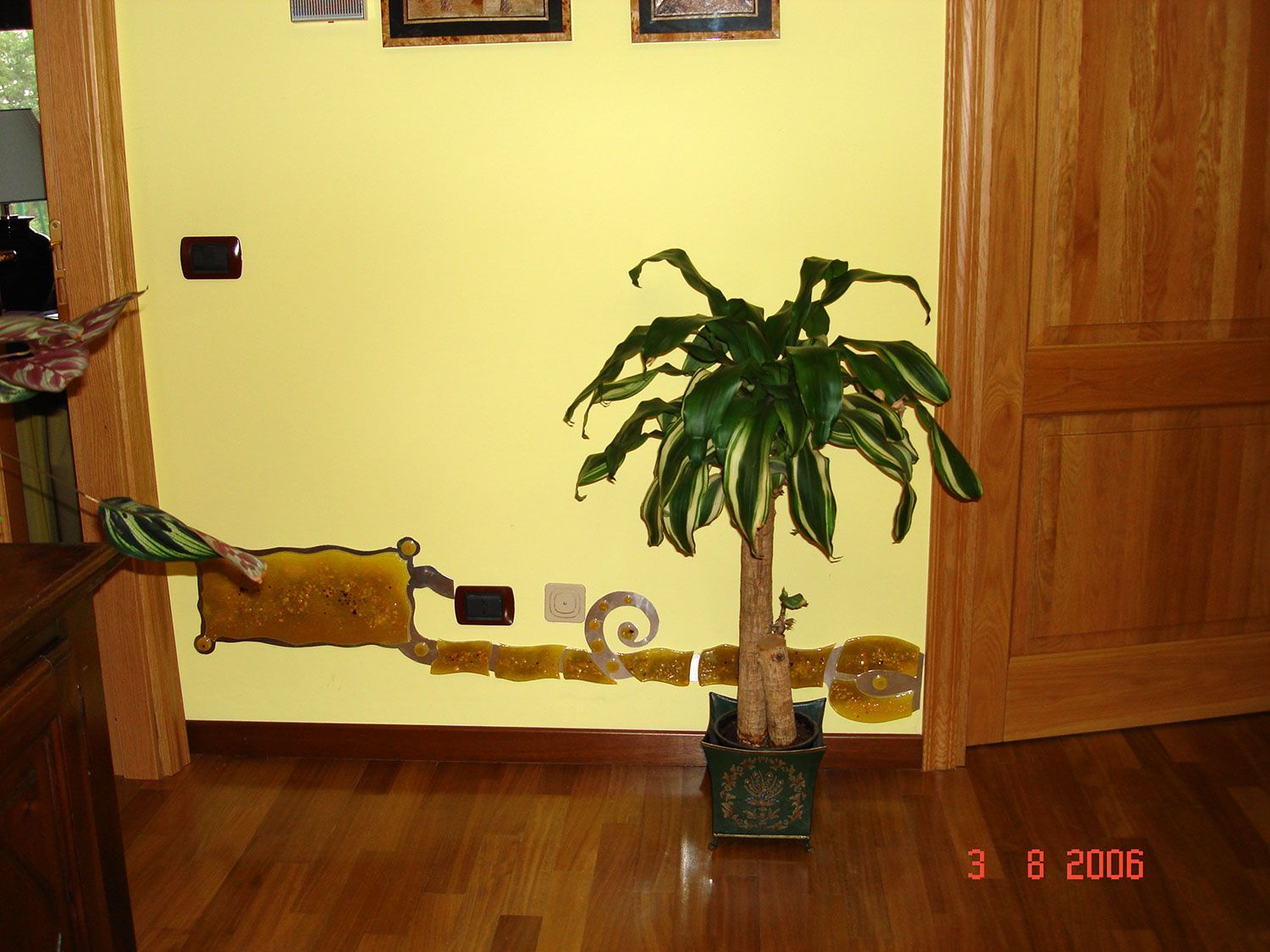 Una pianta di Dracena in un vaso decorativo è appoggiata a una parete gialla con finiture decorative. Pavimento e porta in legno.