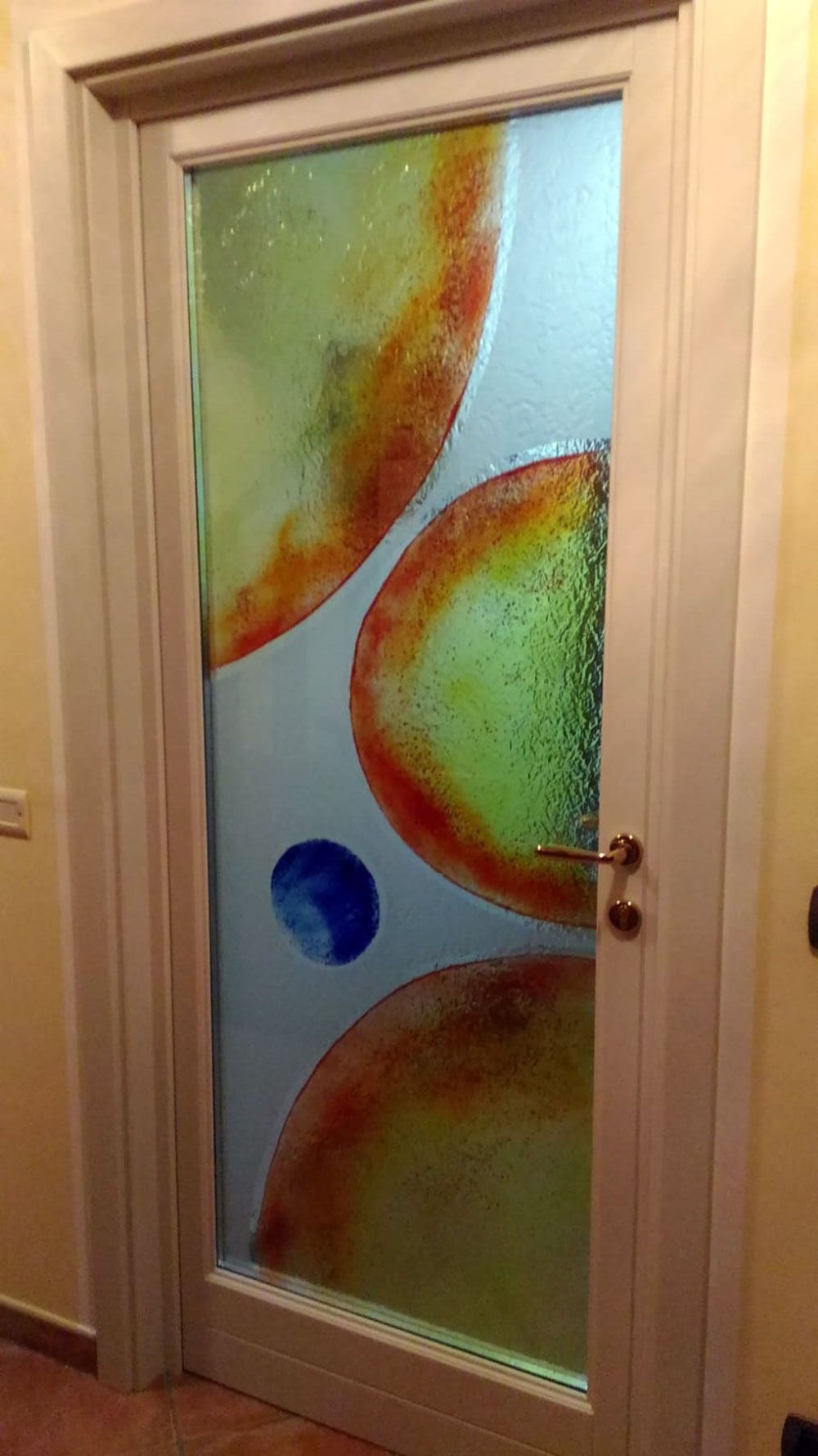 Una porta in vetro con forme astratte colorate in arancione, giallo, verde e blu, inserite in una cornice di colore chiaro.