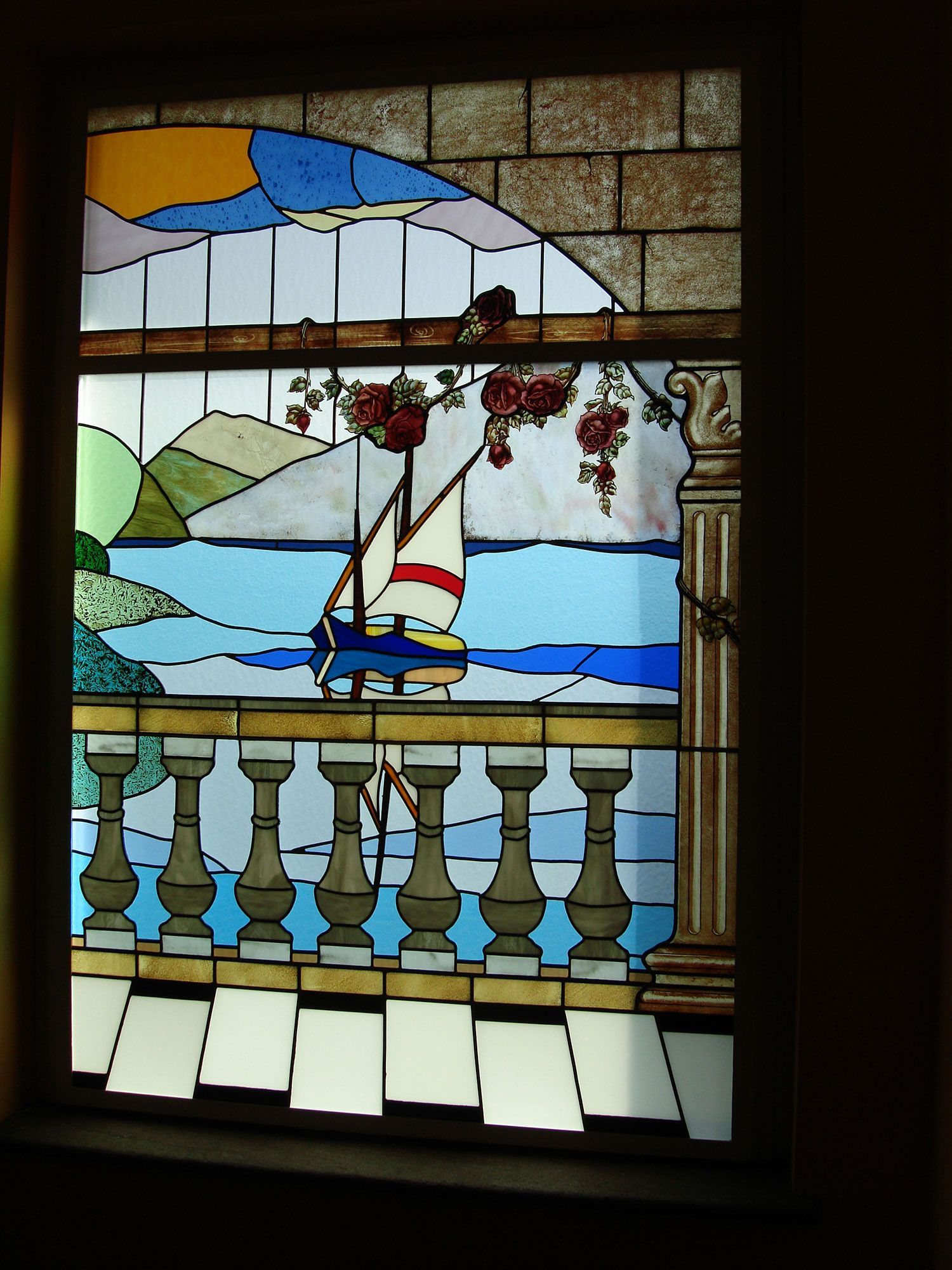 Vetrata con una barca a vela sull'oceano, un balcone e una scena di montagna; vari colori.