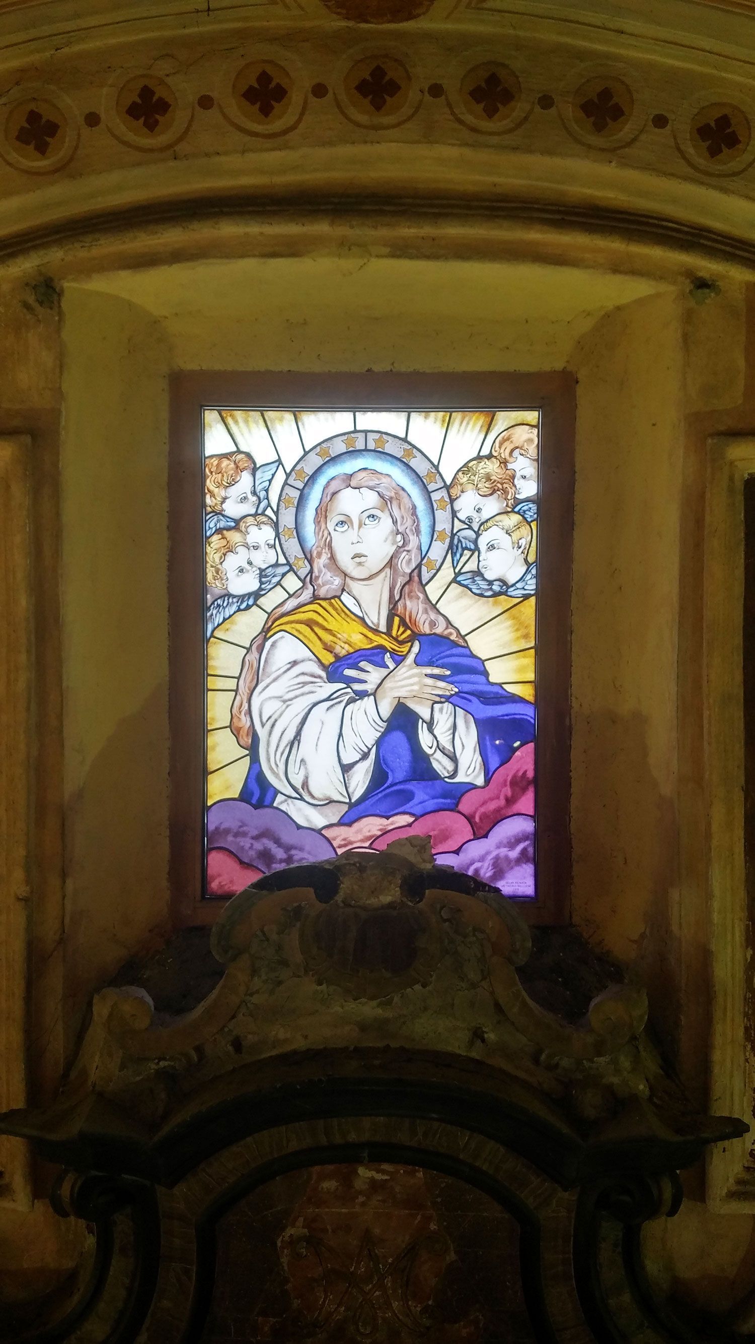 Vetrata: Maria circondata da angeli, vestita di abiti blu e bianchi, in una nicchia ad arco.