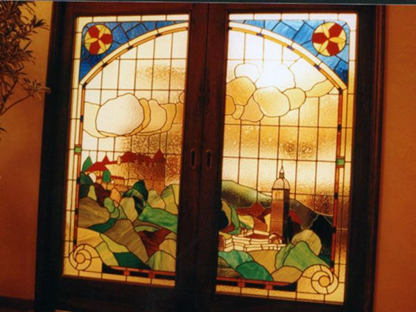 Porte doppie in vetro colorato con una scena paesaggistica: montagne, edifici, cielo e nuvole.