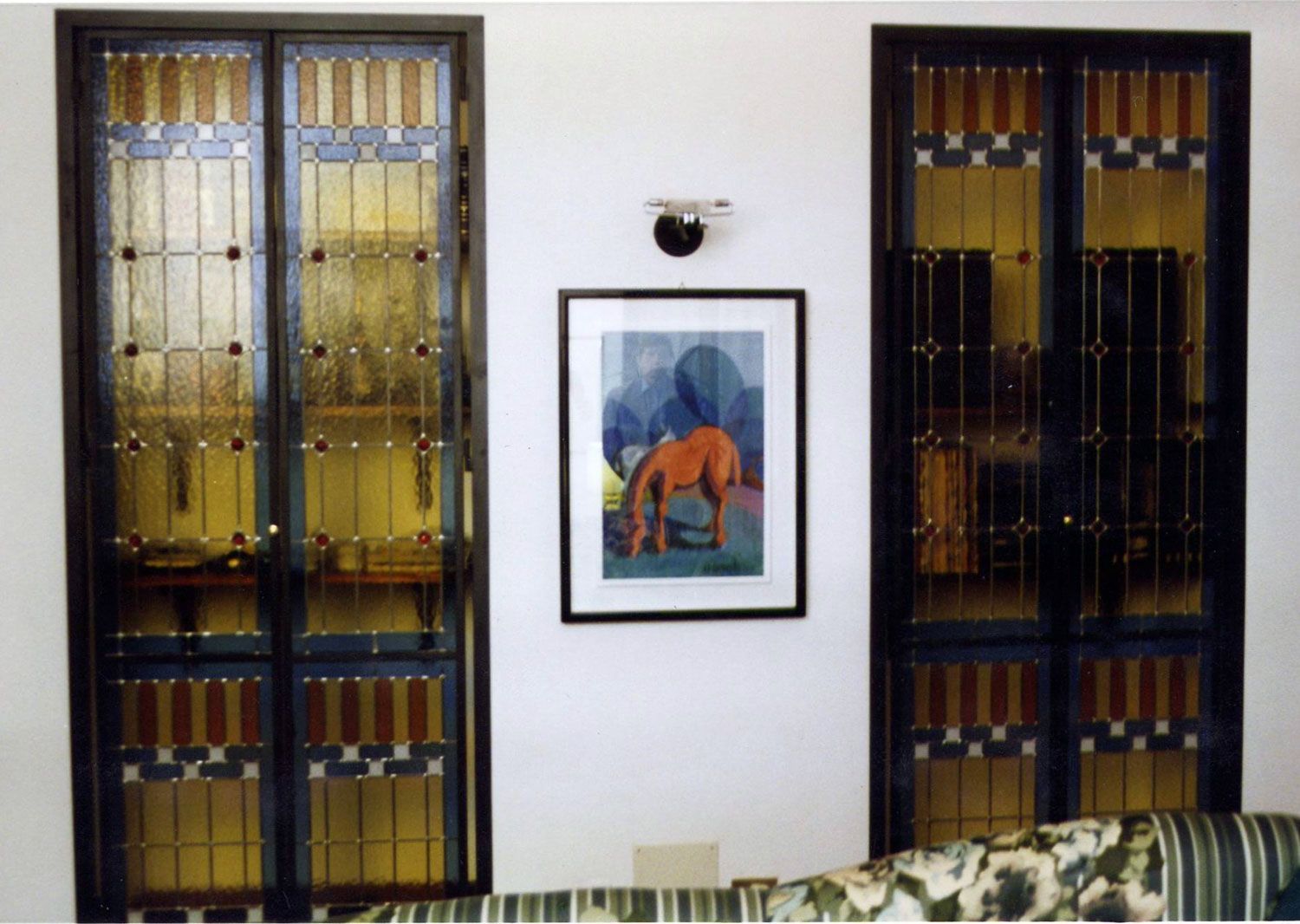 Due porte in vetro colorato fiancheggiano un quadro incorniciato raffigurante un cavallo marrone; parete bianca, lampadario e divano decorato.