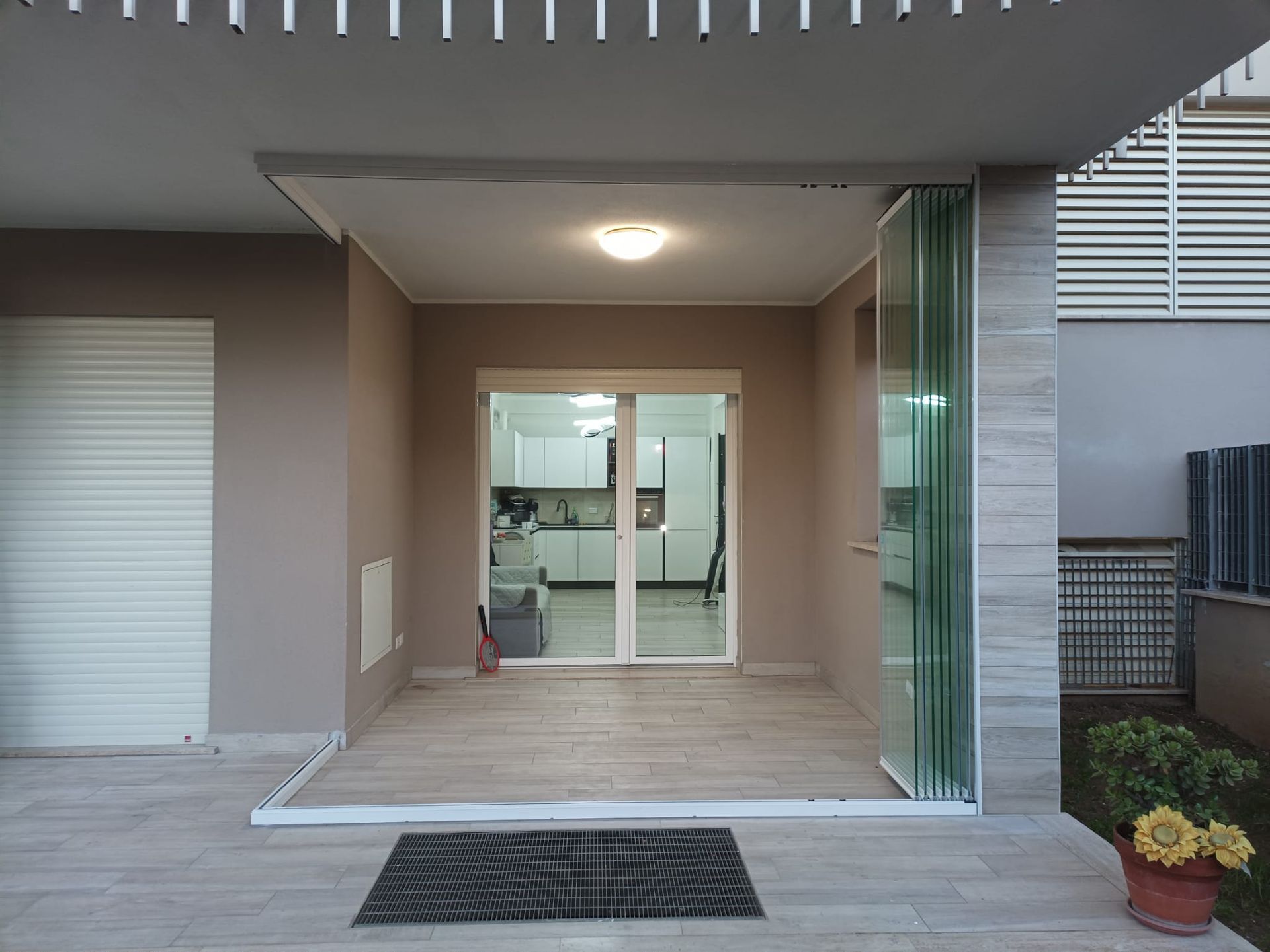 Esterno di un edificio con porte a vetri aperte che conducono alla cucina. Pareti marrone chiaro, pavimento grigio, luce solare.