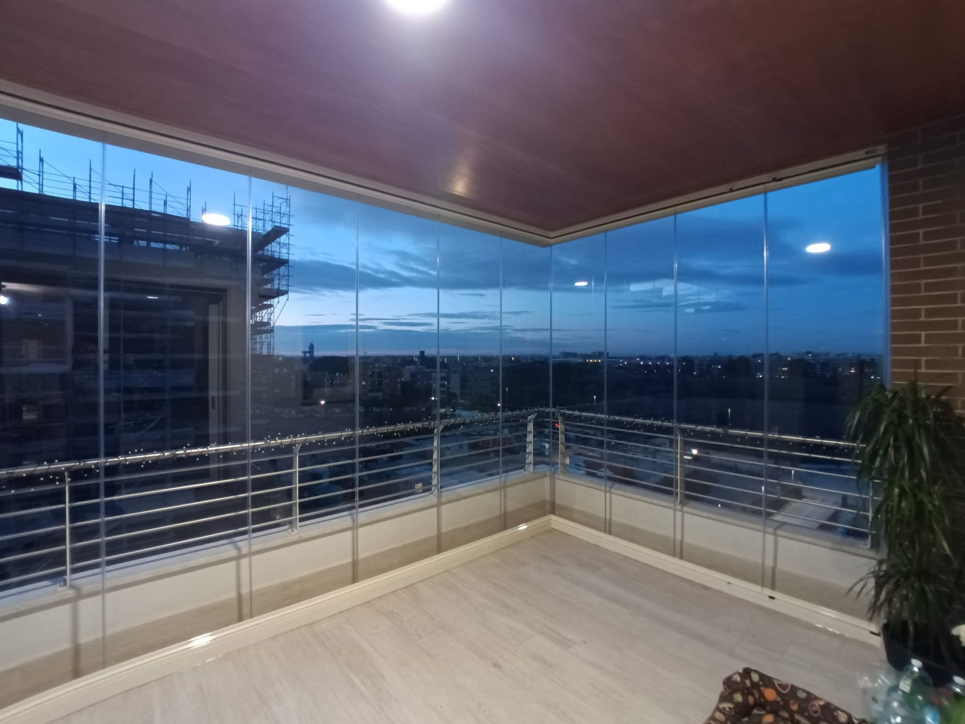 Balcone con pareti in vetro e vista sulla città al tramonto. Pavimento beige, ringhiera in metallo, muro in mattoni e una pianta a vista.