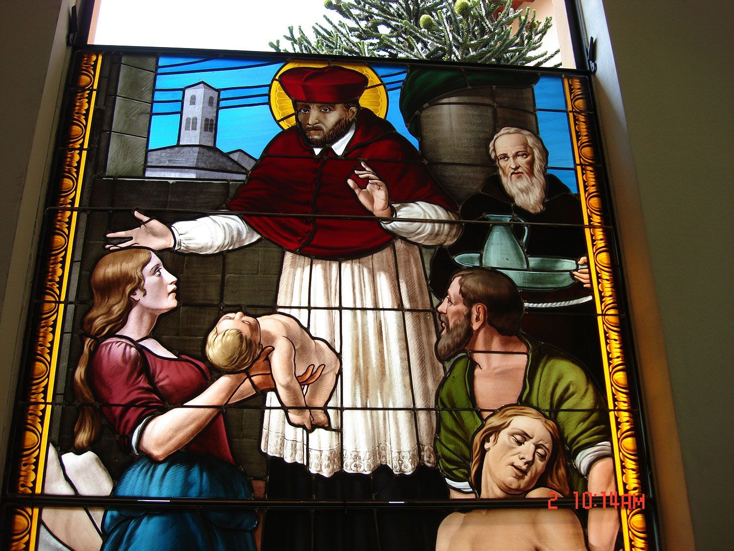 Scena su vetrata: una figura in abito rosso benedice una donna che tiene in braccio un bambino, mentre altri osservano.