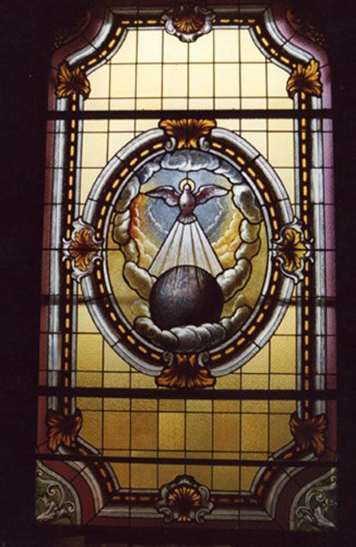 Vetrata: colomba che scende dalle nuvole, irradiando luce su un globo, all'interno di una cornice decorata.