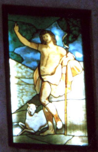 Vetrata raffigurante Gesù con un bastone e un agnello. Alza una mano. Sfumature di blu, giallo e marrone.