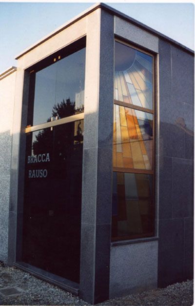 Cripta funeraria con grandi vetrate che riflettono alberi e vetrate colorate. Esterno in granito grigio.