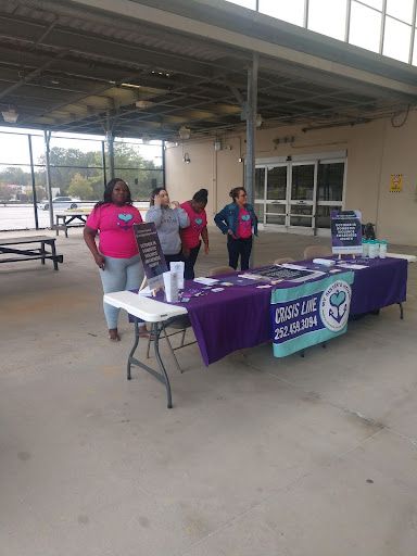 Four women at a table with purple tablecloth promoting Crisis Line. They wear pink shirts. Setting: outdoor, parking area.