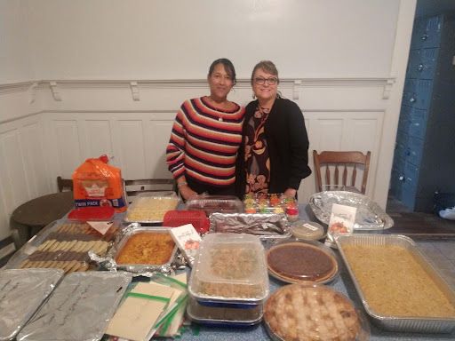 Two women stand behind a table laden with baked goods; indoors, light-colored walls.