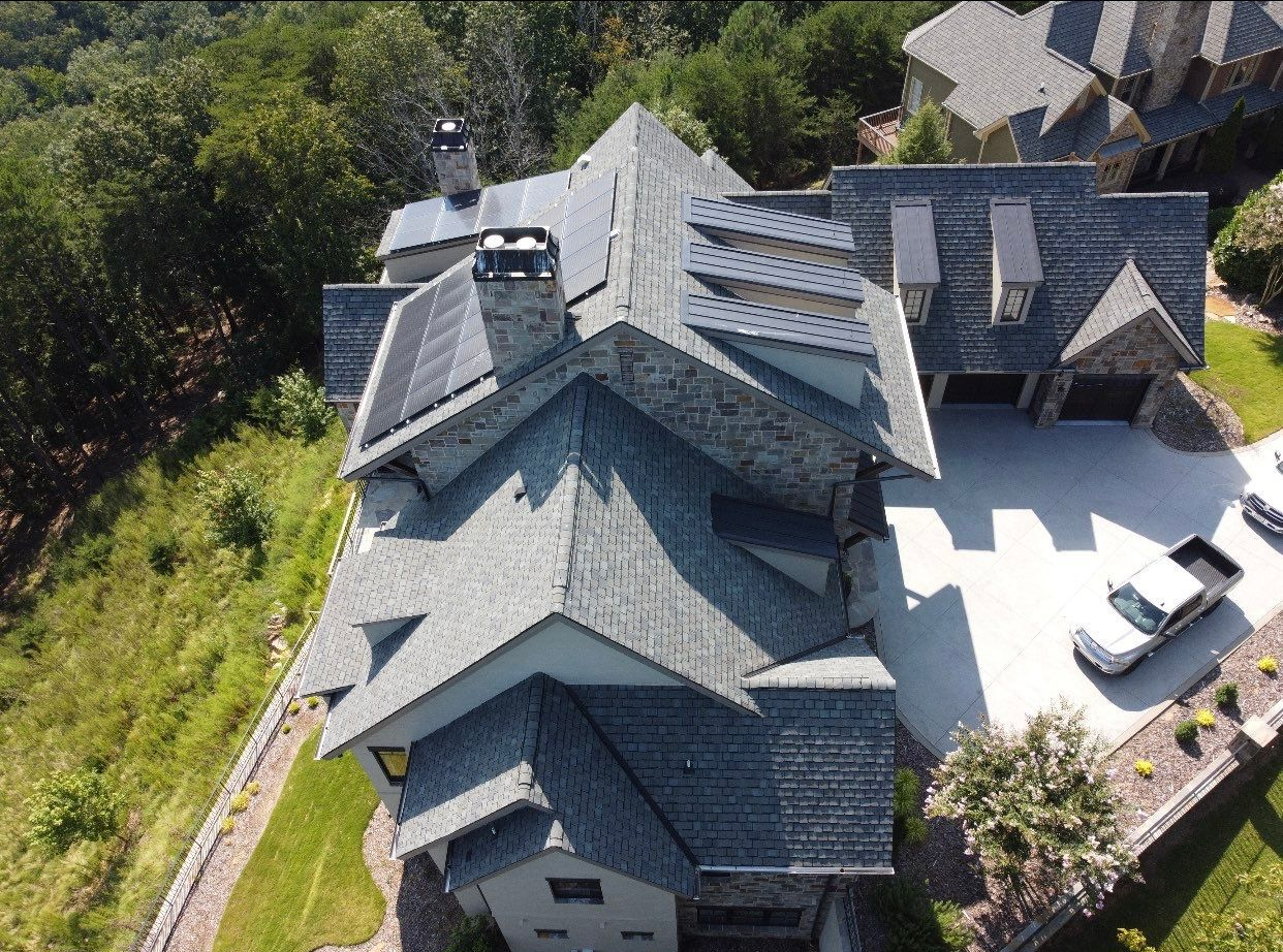 Aerial view of a gray-roofed house with a chimney, driveway, and surrounding greenery.