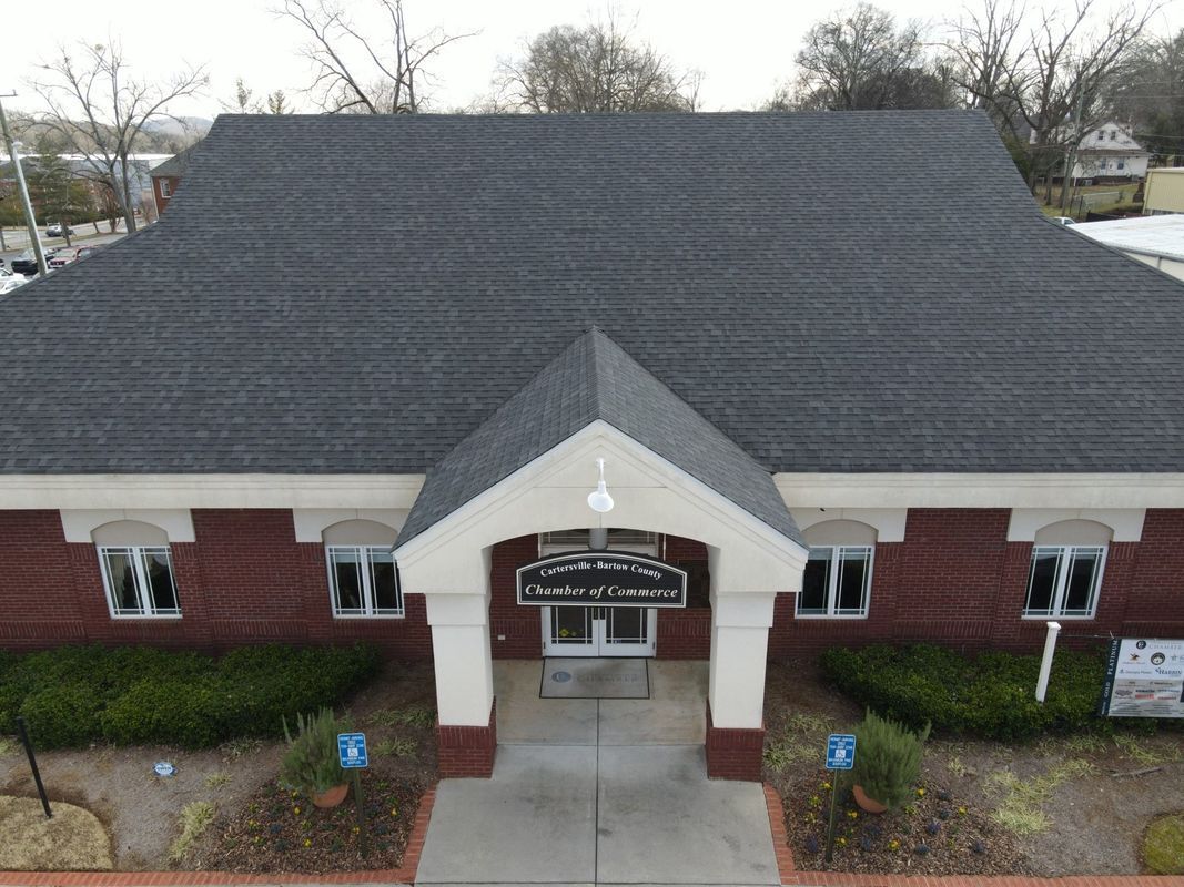 Brick building with dark gray roof, central entrance, windows, and bushes.