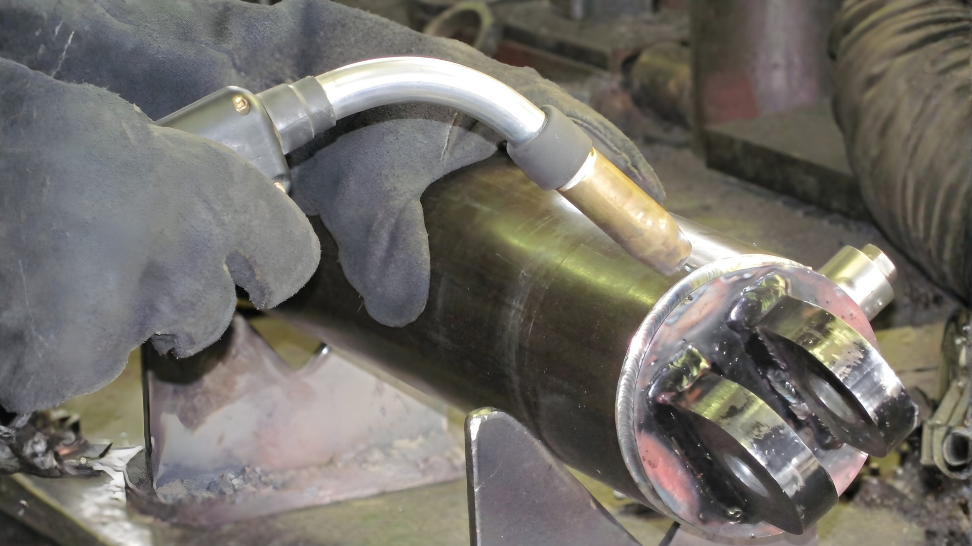 Gloved hands hold a metal torch or tool near a shiny cylindrical pipe in an industrial setting