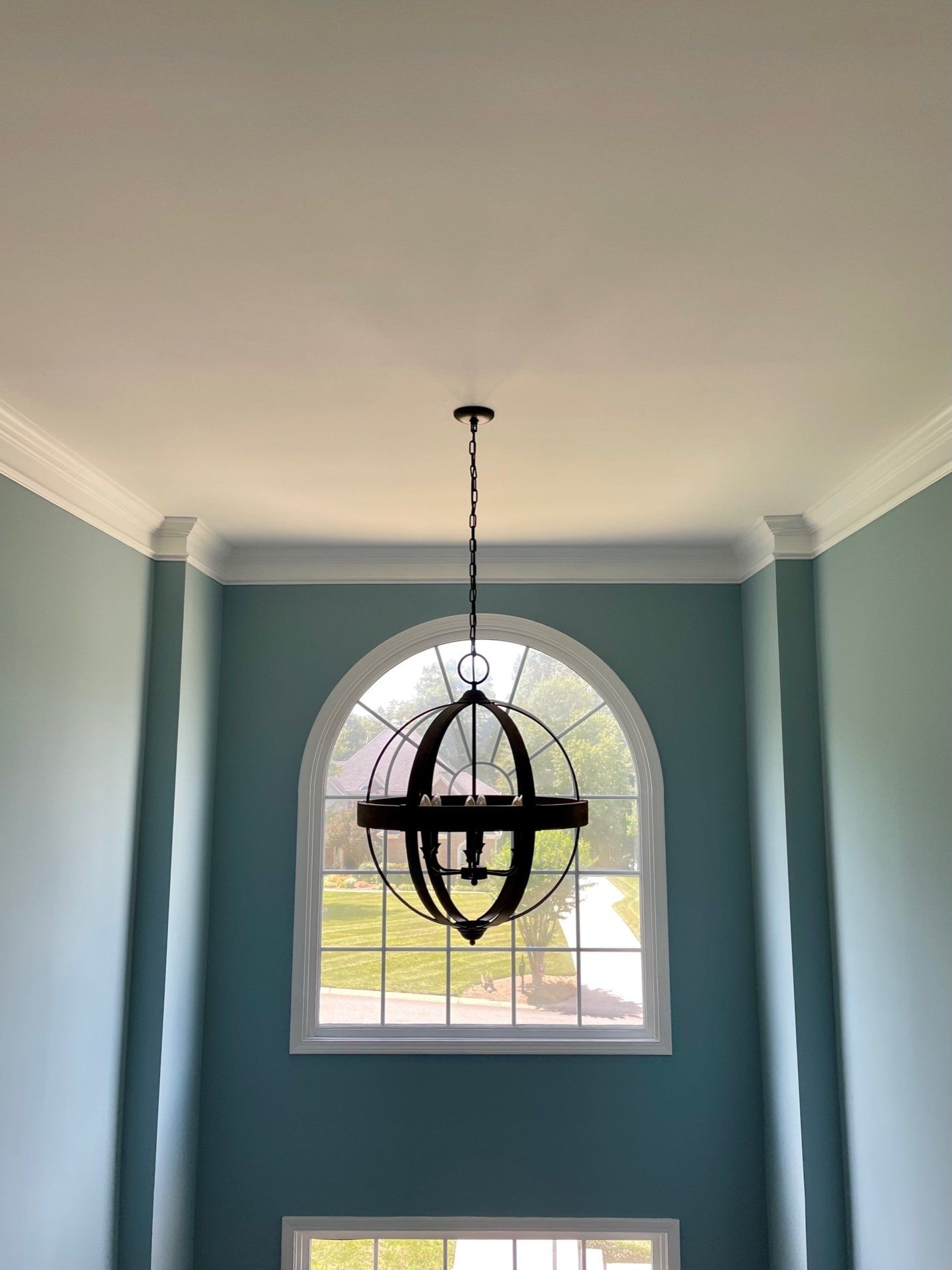 Two-story foyer with teal walls, white trim, and arched window. A dark chandelier hangs in front of the window.