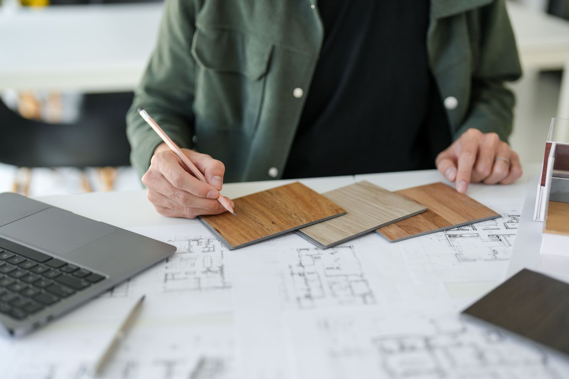 Person drawing on floor plans, selecting wood flooring samples. Laptop, pen and samples on a white desk.