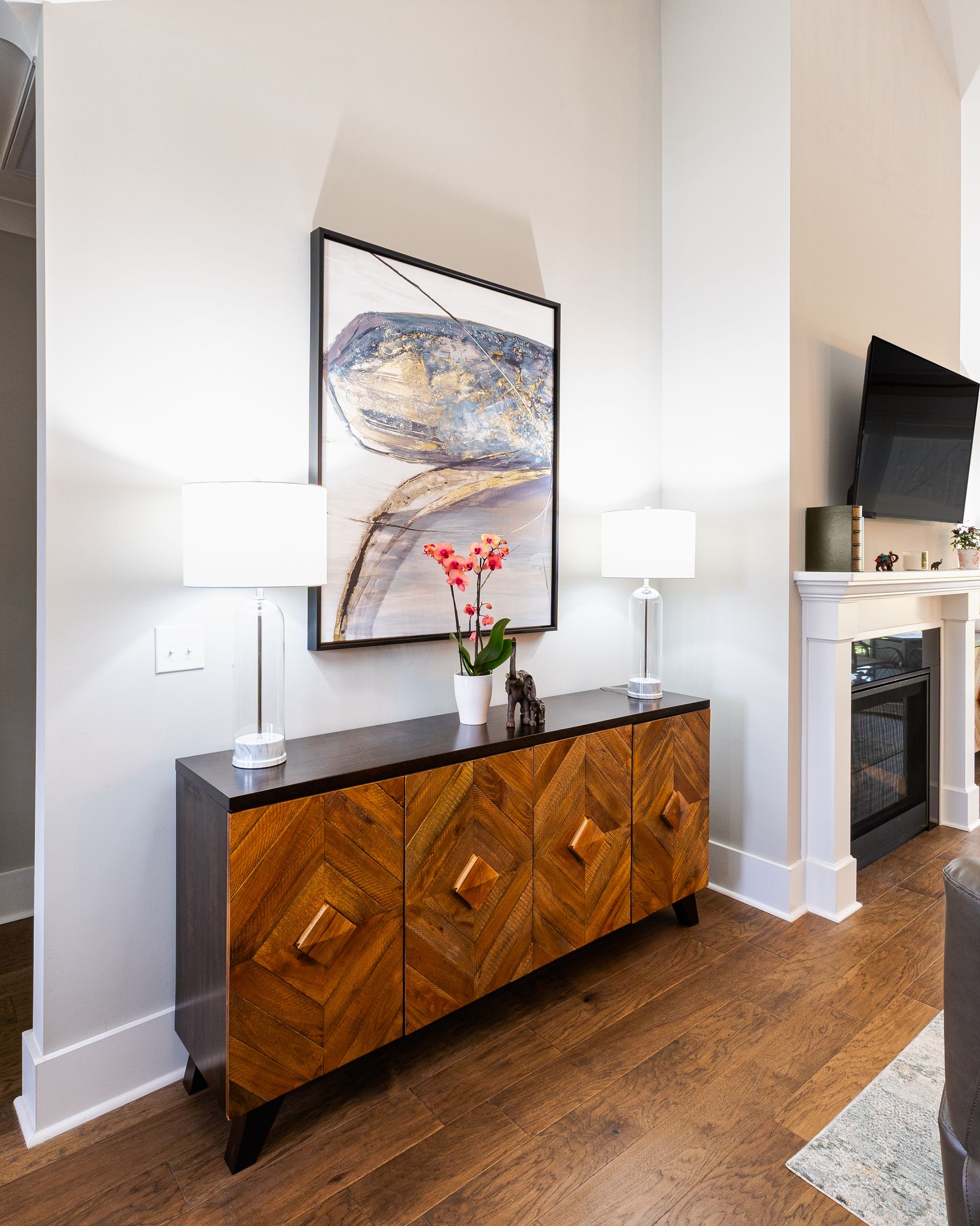 Wooden cabinet with artwork, lamps, and a flower arrangement in a living room.
