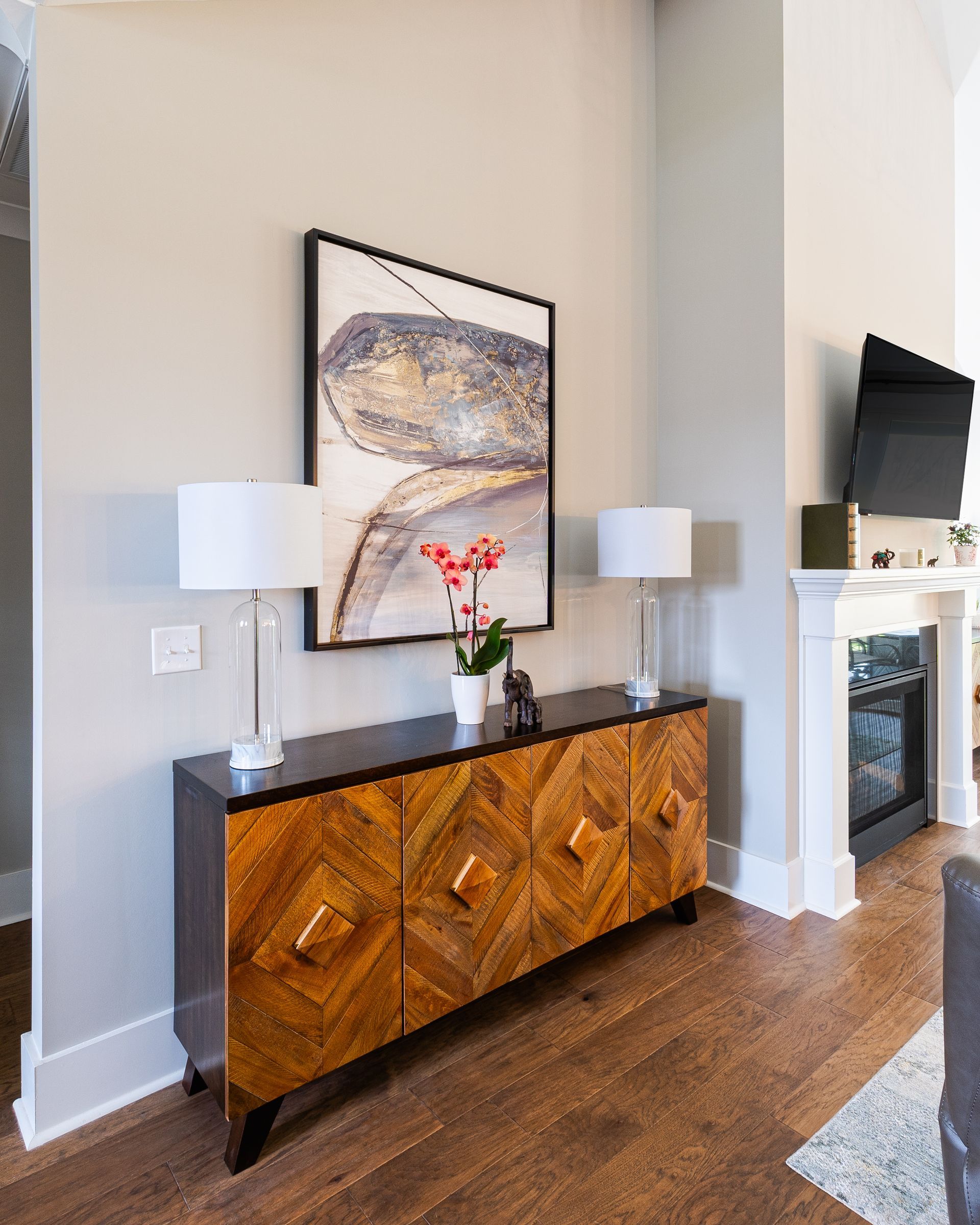 A wooden cabinet with artwork, lamps, and flowers against a light-colored wall, with a fireplace visible.