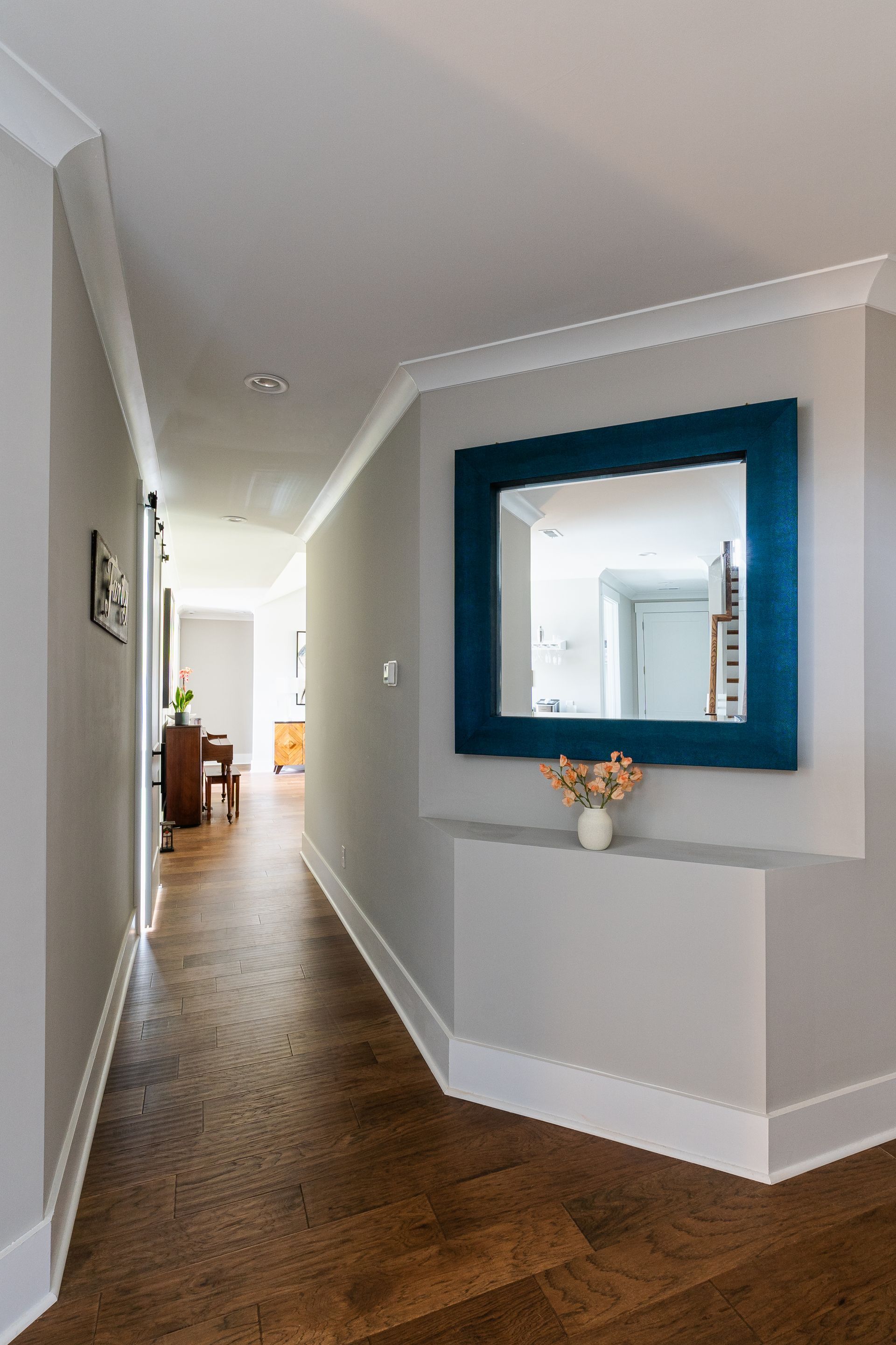 Hallway with a blue-framed mirror above a small ledge with flowers. Dark wood floor, gray walls.