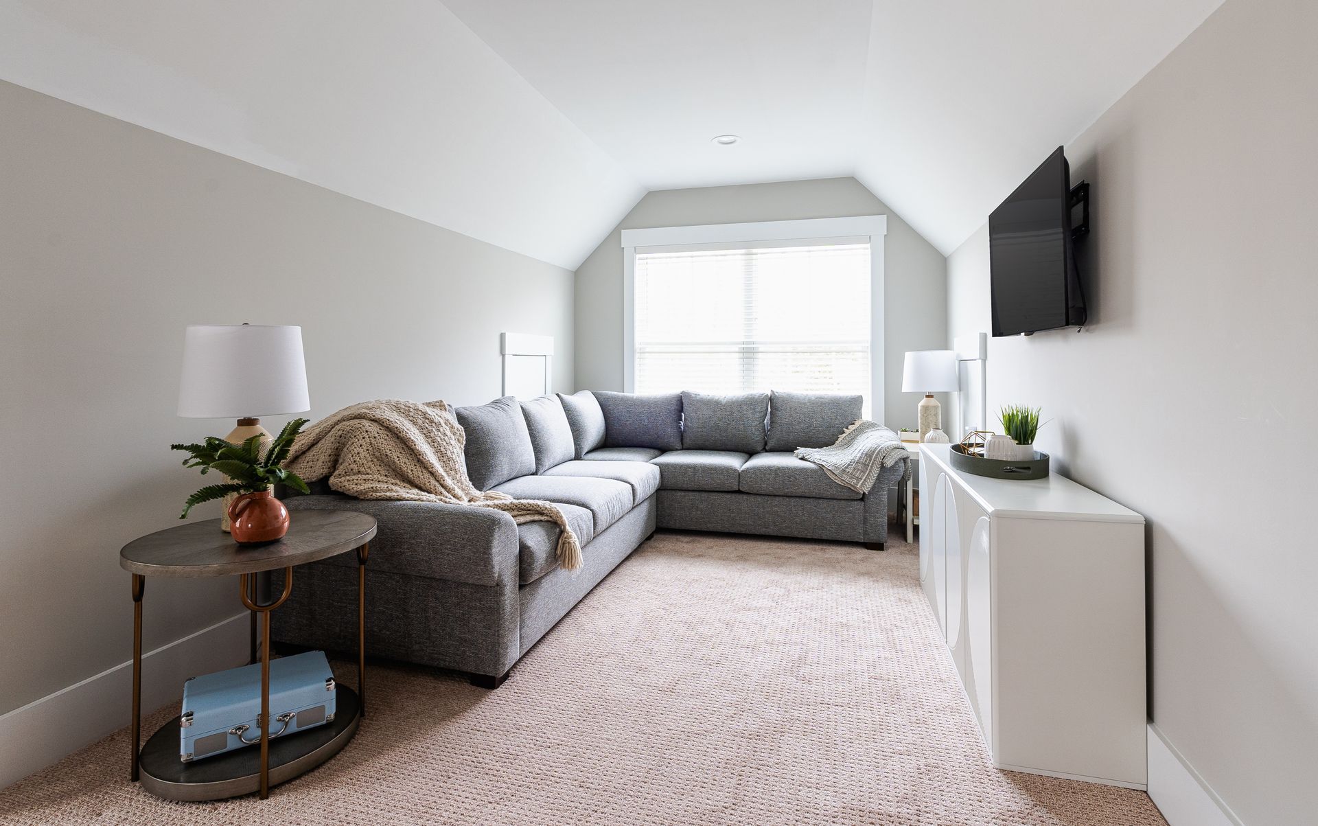Cozy attic living space with gray sectional sofa, pink patterned rug, and white walls. A TV is mounted above a white cabinet.