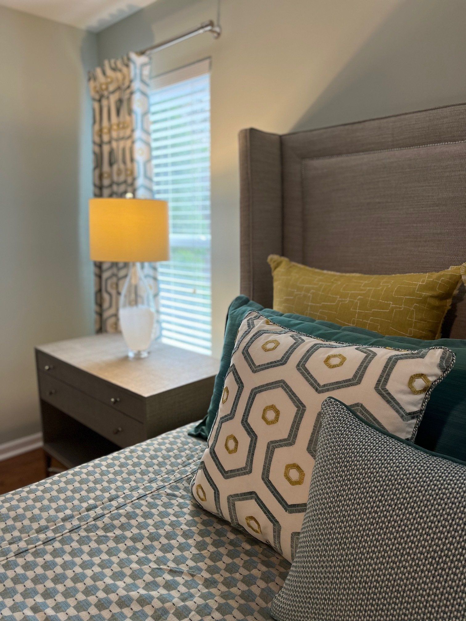 Bedroom with a bed, nightstand, lamp, and decorative pillows. Colors include gray, teal, and yellow.