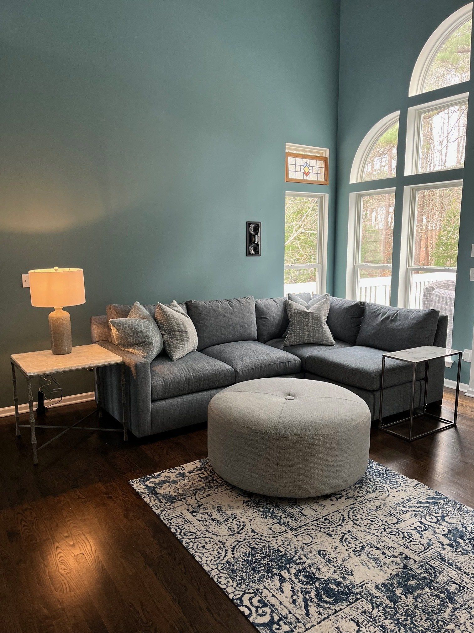 Gray sectional sofa with ottoman and patterned rug in a living room with teal walls and arched windows.